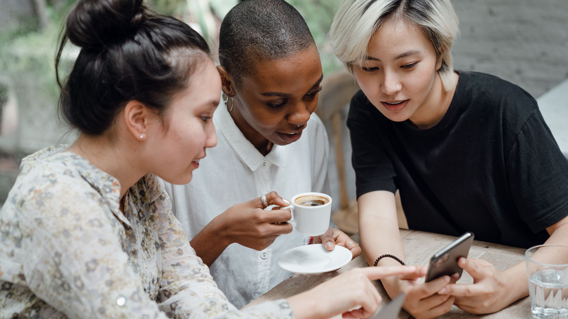 Three people sitting at a table, looking at a smartphone together. One person is holding a coffee cup. They appear to be engaged in a conversation or discussion.